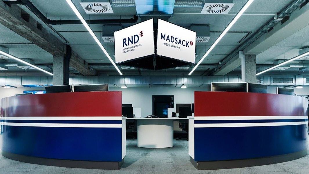 Empfangstresen in Rot-Blau mit h&auml;ngendem RND/MADSACK-Logo-Schild &uuml;ber modernem Redaktionsraum.