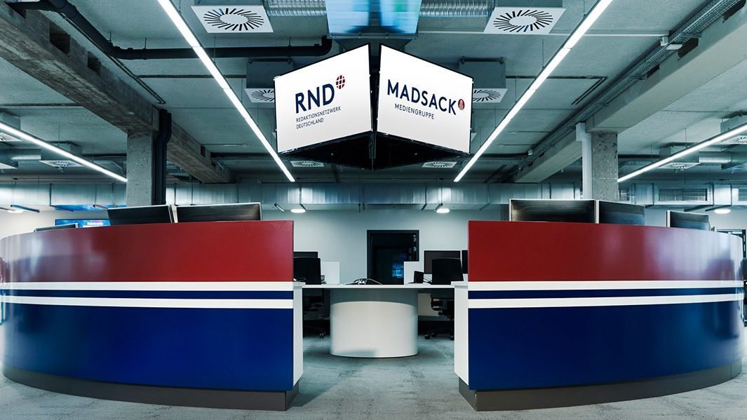 Empfangstresen in Rot-Blau mit h&auml;ngendem RND/MADSACK-Logo-Schild &uuml;ber modernem Redaktionsraum.